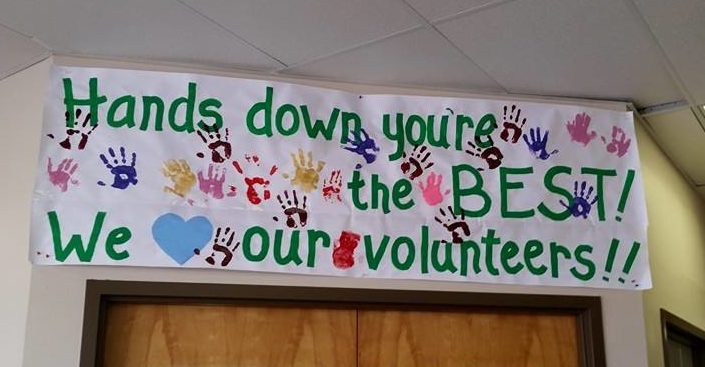 A hand painted signs with multicolored handprints reads, "Hands down you're the BEST! We "heart" our volunteers!"