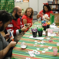 A group of four men and women in christmas attire sit at a table doing crafts. 