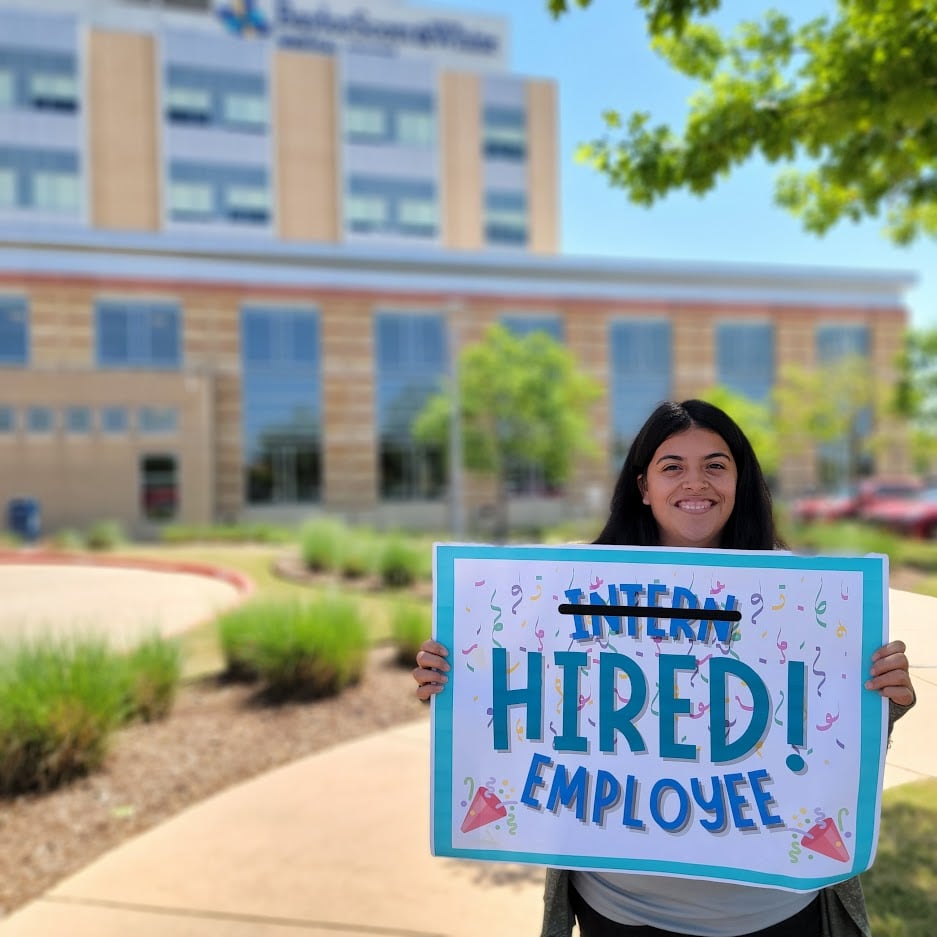 A woman smiles for the camera while holding a sign that reads. "Intern (striked through) HIRED! Employee". In the background is a large hospital building. 