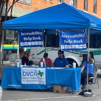BVCIL staff members sit at a table with a blue tablecloth bearing the BVCIL logo under a blue canopy with a sign that reads Accessibility Help Desk. 