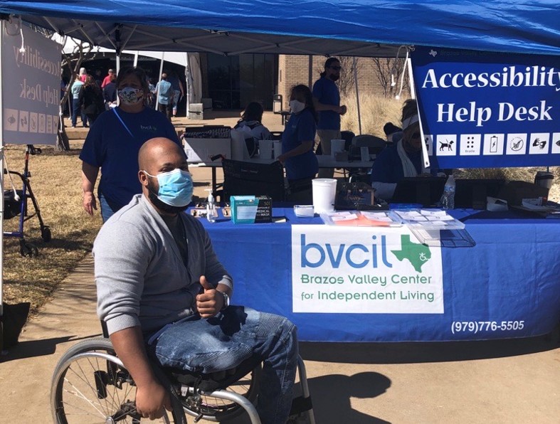 A man sitting in a wheelchair gives a thumbs up to the camera in front of the BVCIL Accessibility Help Desk. 
